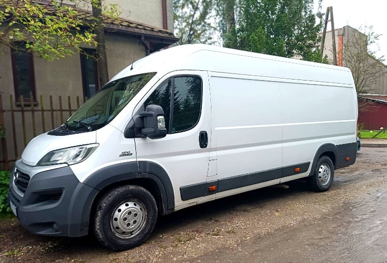 Biały bus dostawczy Fiat Ducato 120 Multijet zaparkowany na mokrej, błotnistej drodze przed budynkiem z zielenią.