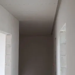 Korytarz w trakcie remontu z zamontowaną płytą gipsowo-kartonową na suficie, widoczne ślady po szpachlowaniu i niezabezpieczone otwory drzwiowe.