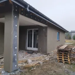 Dom w trakcie ocieplania elewacji: widoczna warstwa izolacji na ścianie, świeżo nałożony tynk na słupie, palety z materiałami budowlanymi na pierwszym planie, pochmurne niebo w tle.