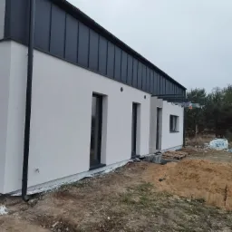 Nowoczesny dom z jasną elewacją i ciemnym metalowym dachem; widoczny fragment elewacji bocznej z oknami i rynną, teren wokół w trakcie prac wykończeniowych, sterta piasku i materiały budowlane w tle.