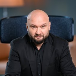 POGORZELSKI NIERUCHOMOŚCI - Portret mężczyzny w czarnym garniturze, z brodą i łysiną, siedzącego na krześle biurowym.