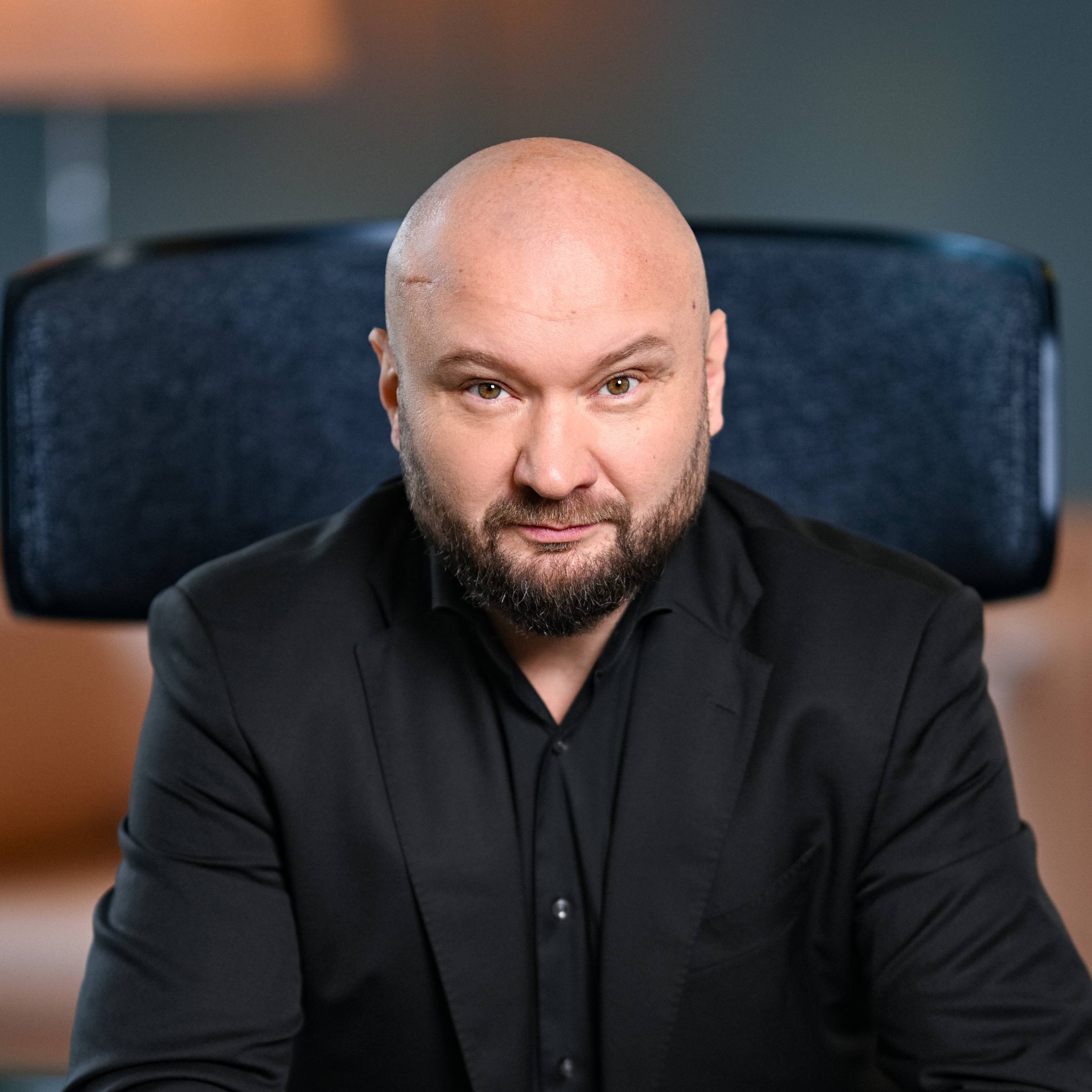 Portret mężczyzny w czarnym garniturze, z brodą i łysiną, siedzącego na krześle biurowym.