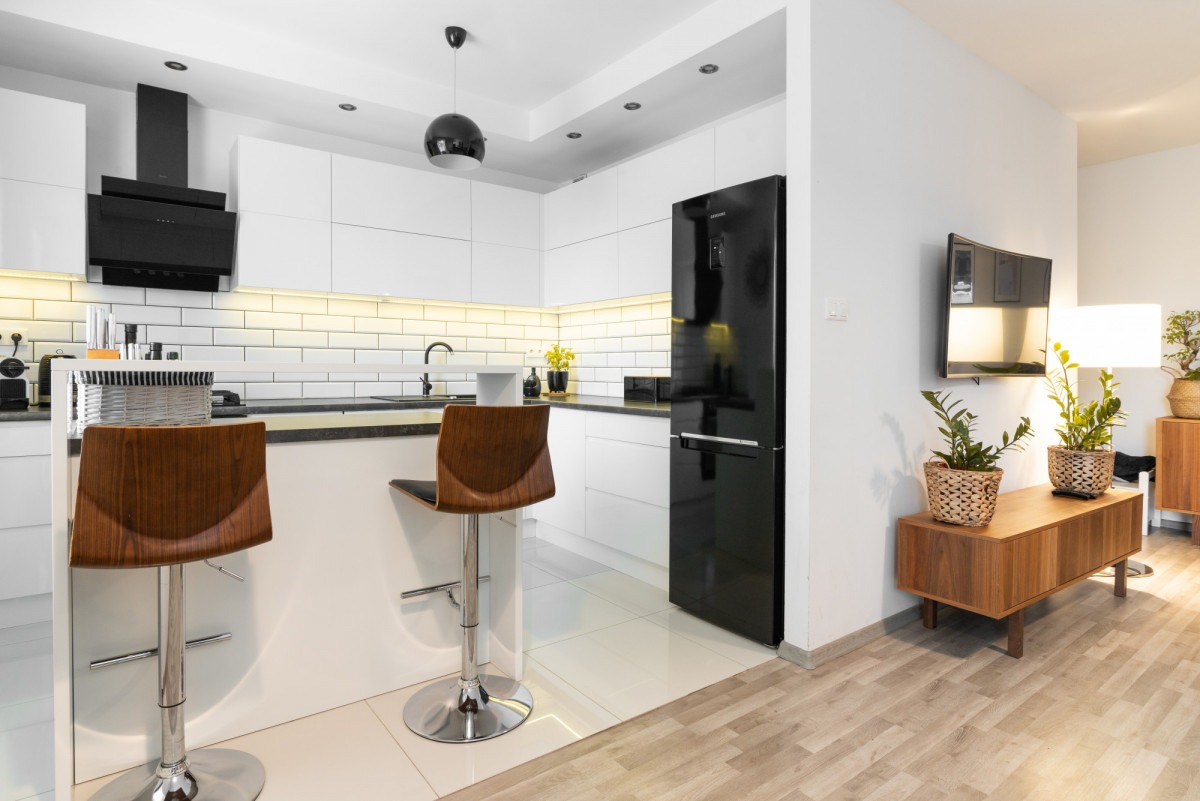 Nowoczesna, jasna kuchnia z białymi szafkami, czarnym sprzętem AGD i drewnianymi hokerami przy wyspie, widok na salon z drewnianą komodą i roślinami doniczkowymi.