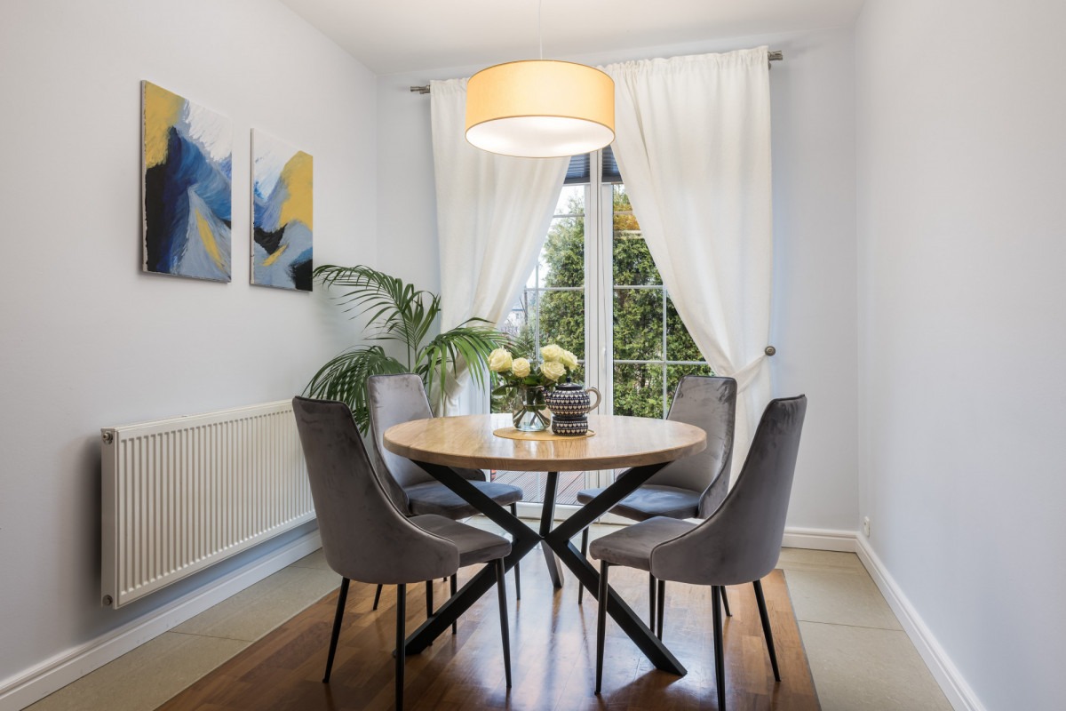 Jasne wnętrze jadalni z okrągłym drewnianym stołem na czarnej, geometrycznej podstawie, czterema szarymi, welurowymi krzesłami, abstrakcyjnymi obrazami na ścianie i rośliną doniczkową w rogu pokoju.