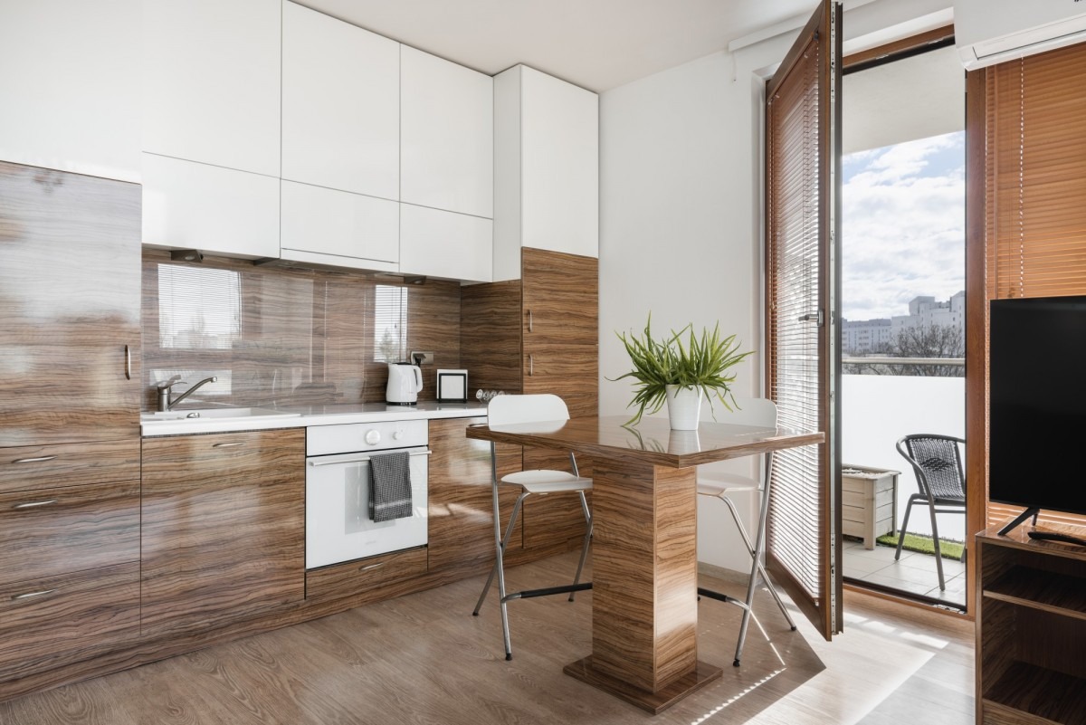 Nowoczesna kuchnia z drewnianymi i białymi szafkami, stołem barowym z rośliną doniczkową, otwartymi drzwiami balkonowymi z żaluzjami i widokiem na miasto w oddali.