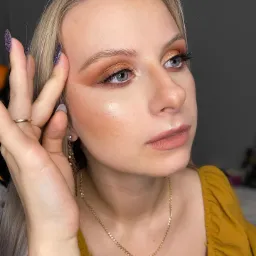 Portret kobiety z blond włosami i złotym cieniem na powiekach, dłonią przy skroni, eksponującą długie, brokatowe paznokcie.