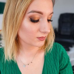 Beauty Center - Portret kobiety z profesjonalnym, błyszczącym makijażem oczu w odcieniach złota i brązu, wykonanym w Nowym Targu. Widoczne sztuczne rzęsy i delikatny makijaż ust. Kobieta ma blond włosy i zielony...