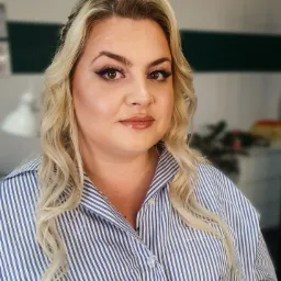 Portret kobiety z profesjonalnym makijażem, widoczne mocno podkreślone oczy eyelinerem, błyszczące usta i delikatny kontur twarzy, blond włosy ułożone w fale, ubrana w koszulę w niebiesko-białe paski.