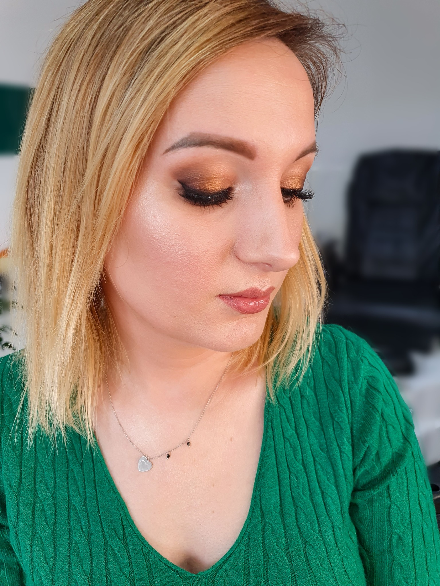 Portret kobiety z profesjonalnym, błyszczącym makijażem oczu w odcieniach złota i brązu, wykonanym w Nowym Targu. Widoczne sztuczne rzęsy i delikatny makijaż ust. Kobieta ma blond włosy i zielony...