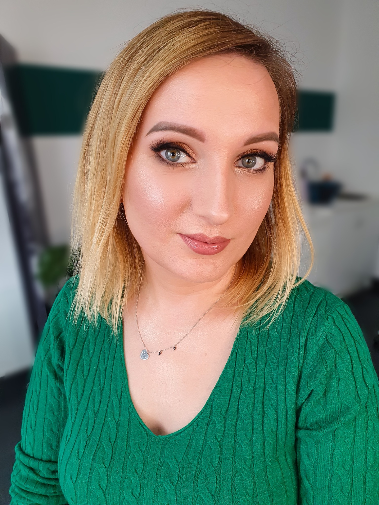 Portret kobiety z blond włosami w zielonym swetrze, z delikatnym makijażem oczu i błyszczącymi ustami, patrzącej w obiektyw.