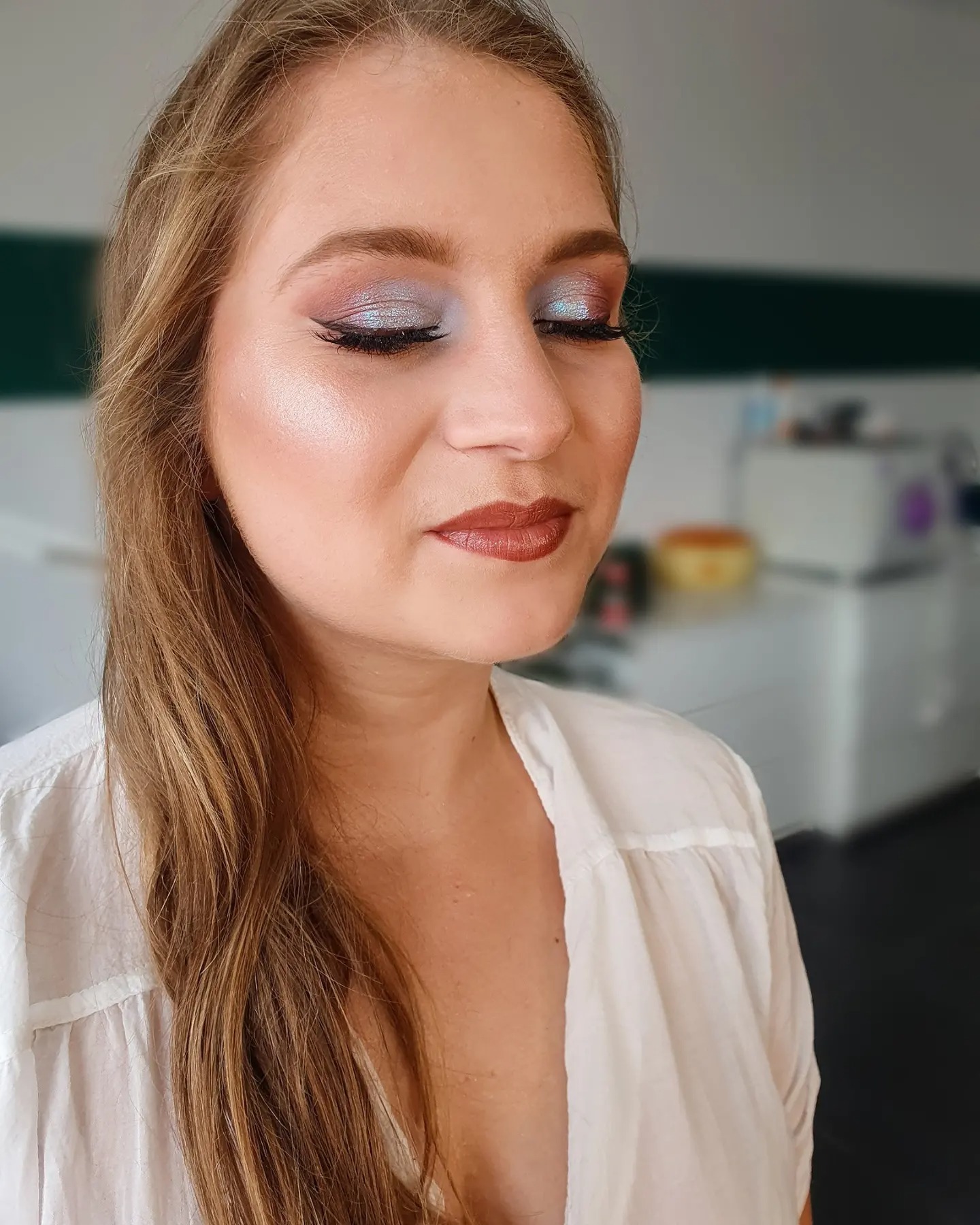 Portret kobiety z zamkniętymi oczami, prezentującej srebrzysto-niebieski makijaż oczu z czarną kreską i błyszczącą, brązową pomadką. Widoczny kontur twarzy i rozświetlona skóra.