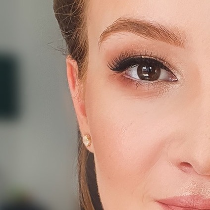 Detal portretu kobiety z delikatnym makijażem oczu w odcieniach brązu i beżu, z wyraźnie podkreślonymi rzęsami i błyszczącą złotą kolczykiem w uchu.