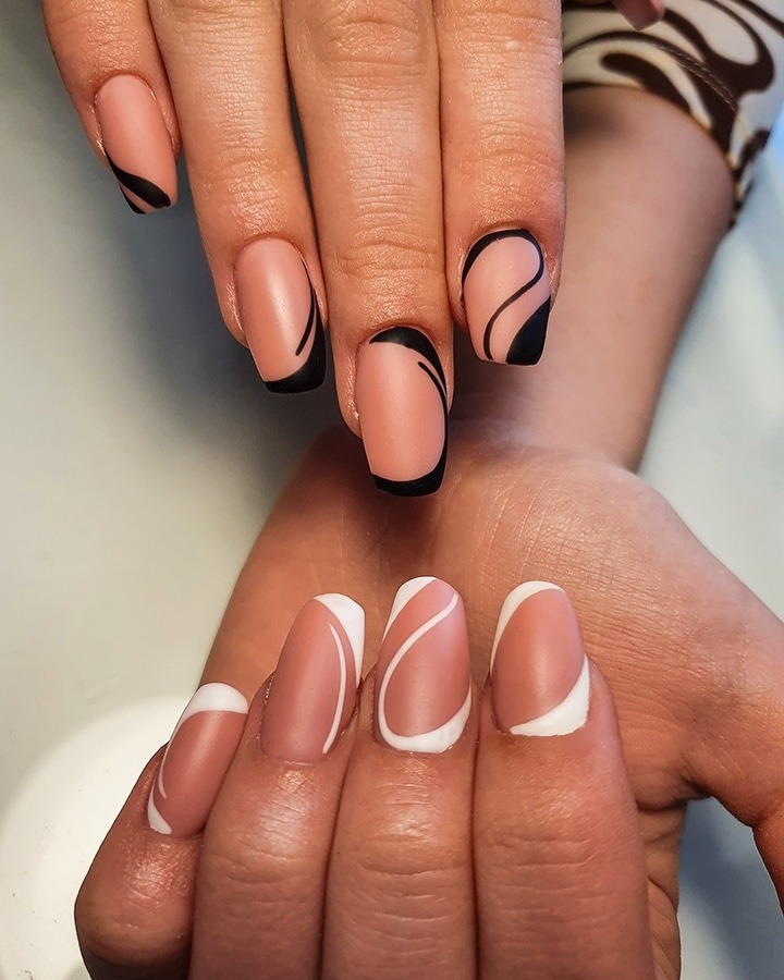 Dłonie z pomalowanymi paznokciami w geometryczne wzory: czarne fale na beżowym tle na jednej dłoni i białe fale na beżowym tle na drugiej dłoni. Matowe wykończenie.