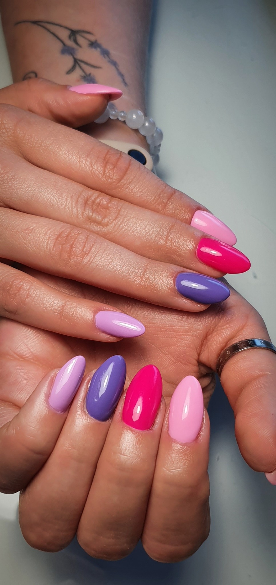Zbliżenie dłoni z pomalowanymi paznokciami w odcieniach różu i fioletu, migdałowy kształt, pierścionek na palcu wskazującym, bransoletka z koralików i tatuaż lawendy na nadgarstku.