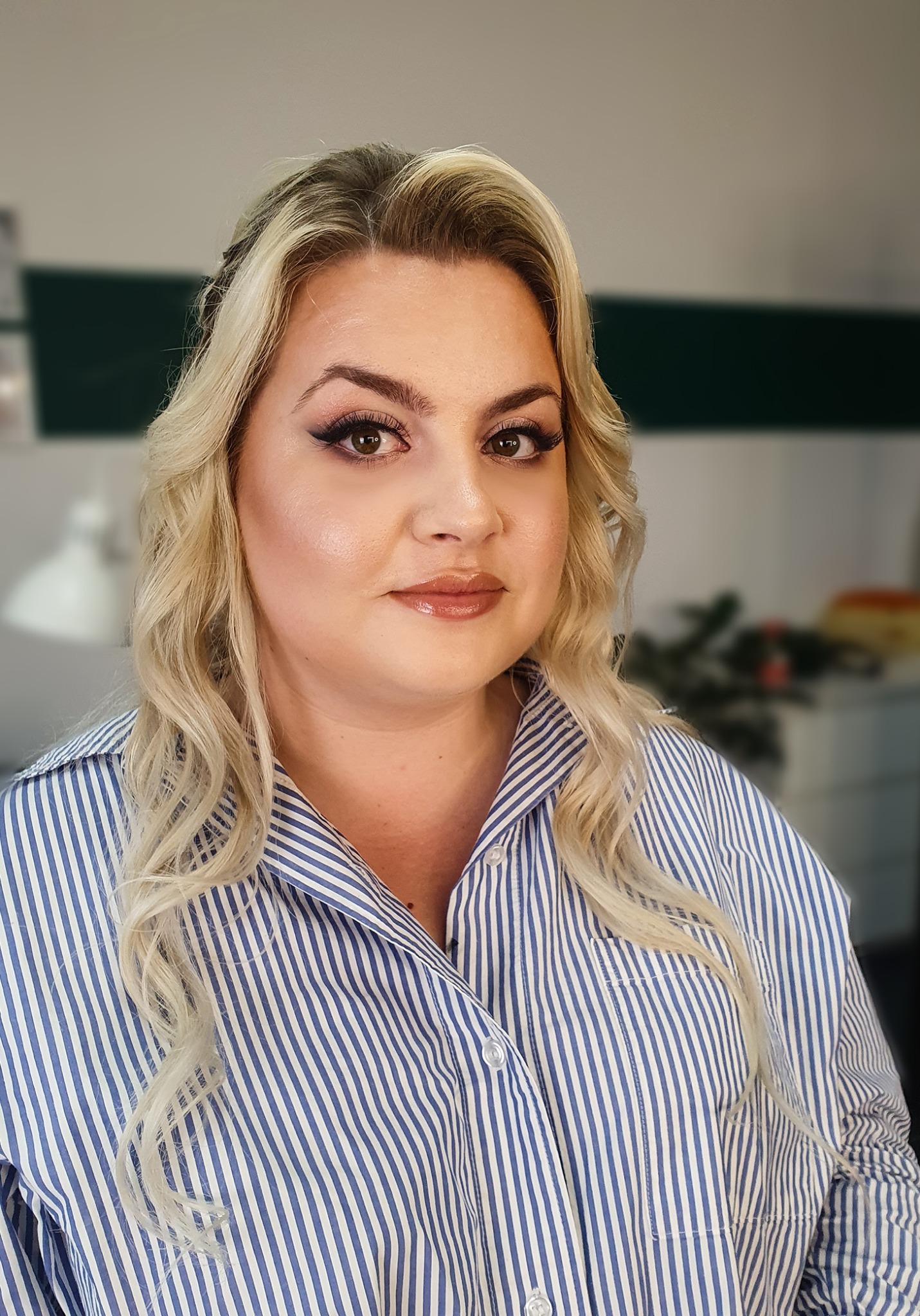 Portret kobiety z profesjonalnym makijażem, widoczne mocno podkreślone oczy eyelinerem, błyszczące usta i delikatny kontur twarzy, blond włosy ułożone w fale, ubrana w koszulę w niebiesko-białe paski.
