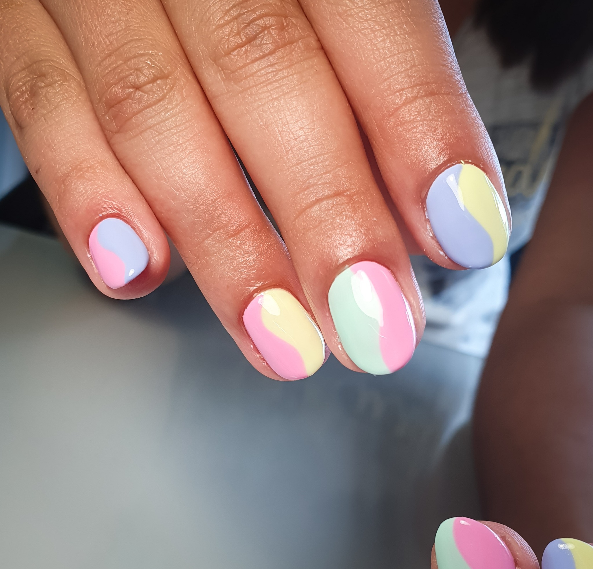 Dłoń z pomalowanymi paznokciami w pastelowych kolorach: różowym, fioletowym, żółtym i miętowym, tworzącymi wzór fali na każdym paznokciu.