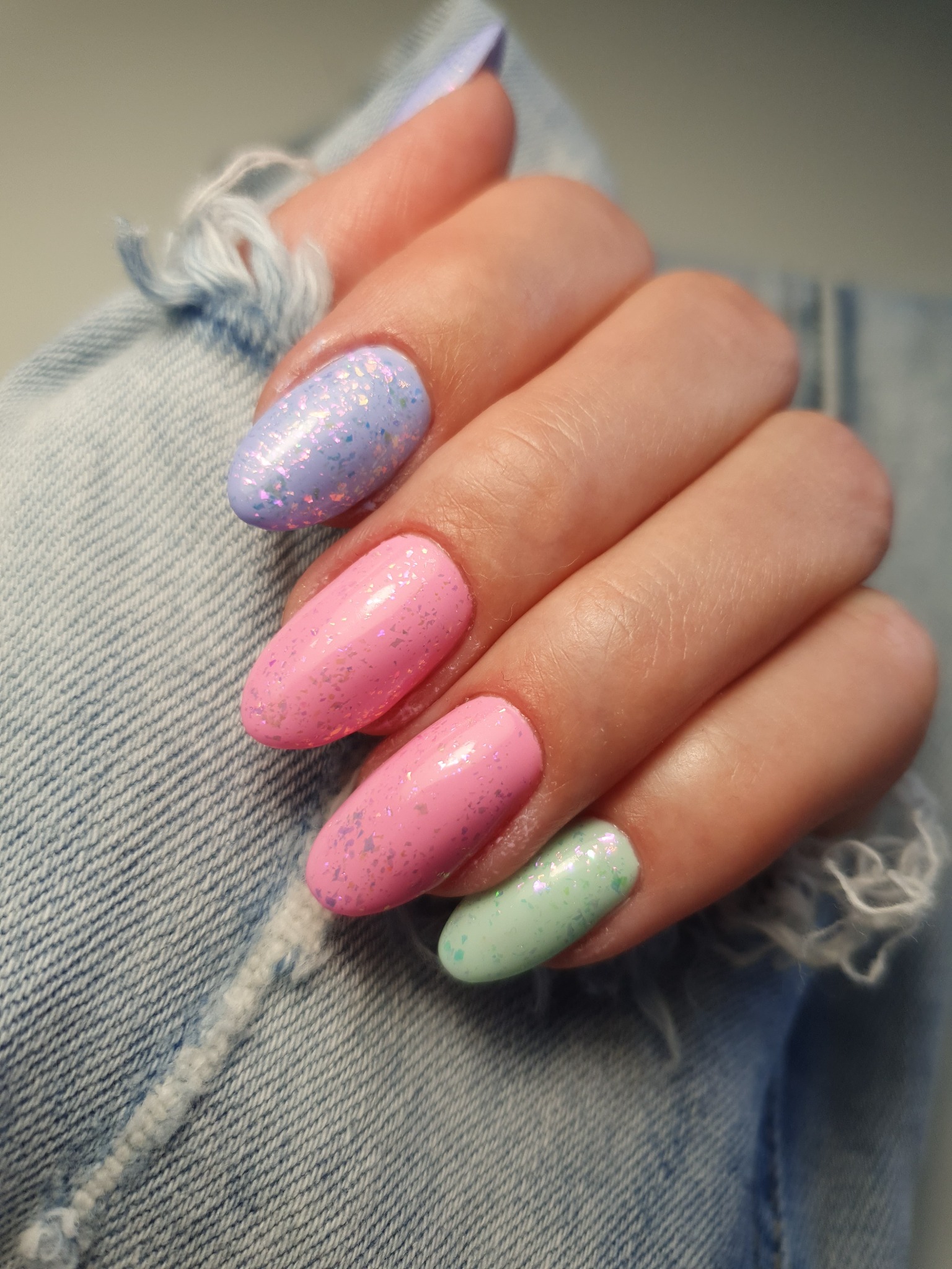 Dłoń z pastelowymi paznokciami w odcieniach różu, błękitu i zieleni, ozdobionymi brokatem, na tle podartego denimu.