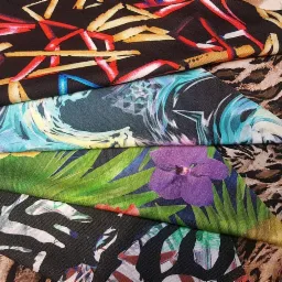 Kolekcja próbek tkanin z różnorodnymi wzorami: abstrakcyjne pociągnięcia pędzlem, motywy fal, tropikalne liście z fioletowym kwiatem oraz wzór zebry i panterki.