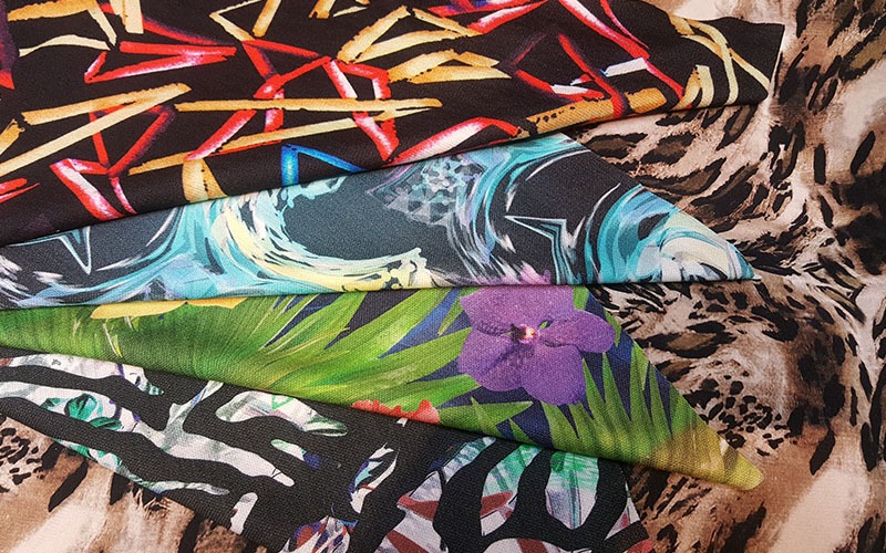 Kolekcja próbek tkanin z różnorodnymi wzorami: abstrakcyjne pociągnięcia pędzlem, motywy fal, tropikalne liście z fioletowym kwiatem oraz wzór zebry i panterki.