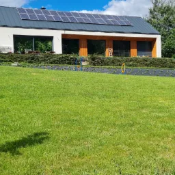 Nowoczesny dom z panelami słonecznymi na dachu, widok z zadbanego trawnika z młodymi drzewkami i przygotowaną rabatą z rozłożoną agrowłókniną i nasadzeniami.