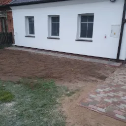 Świeżo otynkowana, biała ściana domu z trzema oknami i nowym dachem z ciemnej dachówki, obok ceglany budynek i przygotowany teren pod trawnik oraz wyłożona kostką ścieżka.