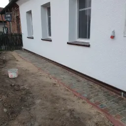 Nowo położona kostka brukowa wzdłuż ściany budynku z oknami, z widocznym wiaderkiem i ziemią po bokach.