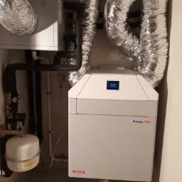 Zamknięta instalacja rekuperacji z centralą Aventa PRO AUROS 605, rury wentylacyjne w srebrnej izolacji, czarne rury hydrauliczne i biały zbiornik wyrównawczy na tle jasnej ściany.