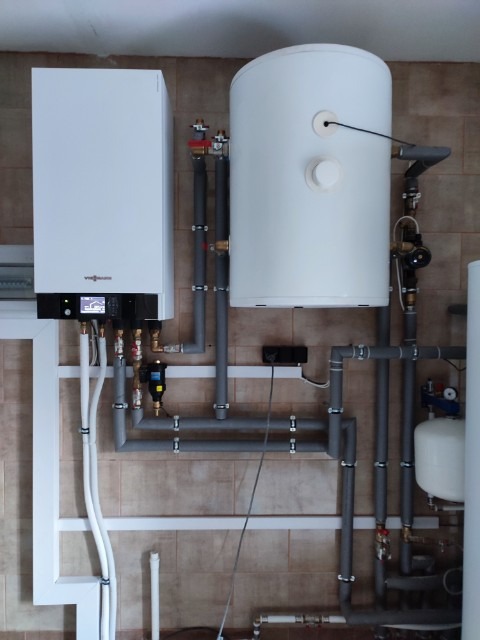 Biały, wiszący kocioł grzewczy Viessmann z podłączonym zasobnikiem ciepłej wody i instalacją hydrauliczną z szarych rur PVC, zamontowany na tle beżowych płytek ceramicznych.