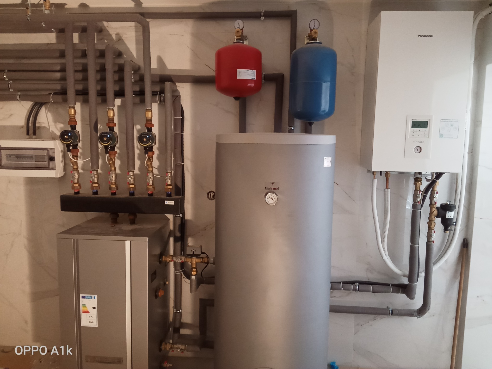 Szara kotłownia z białym kotłem gazowym Panasonic, szarym zbiornikiem Galmet, czerwonym i niebieskim naczyniem przeponowym oraz rurami w izolacji, całość na tle marmurowej ściany.