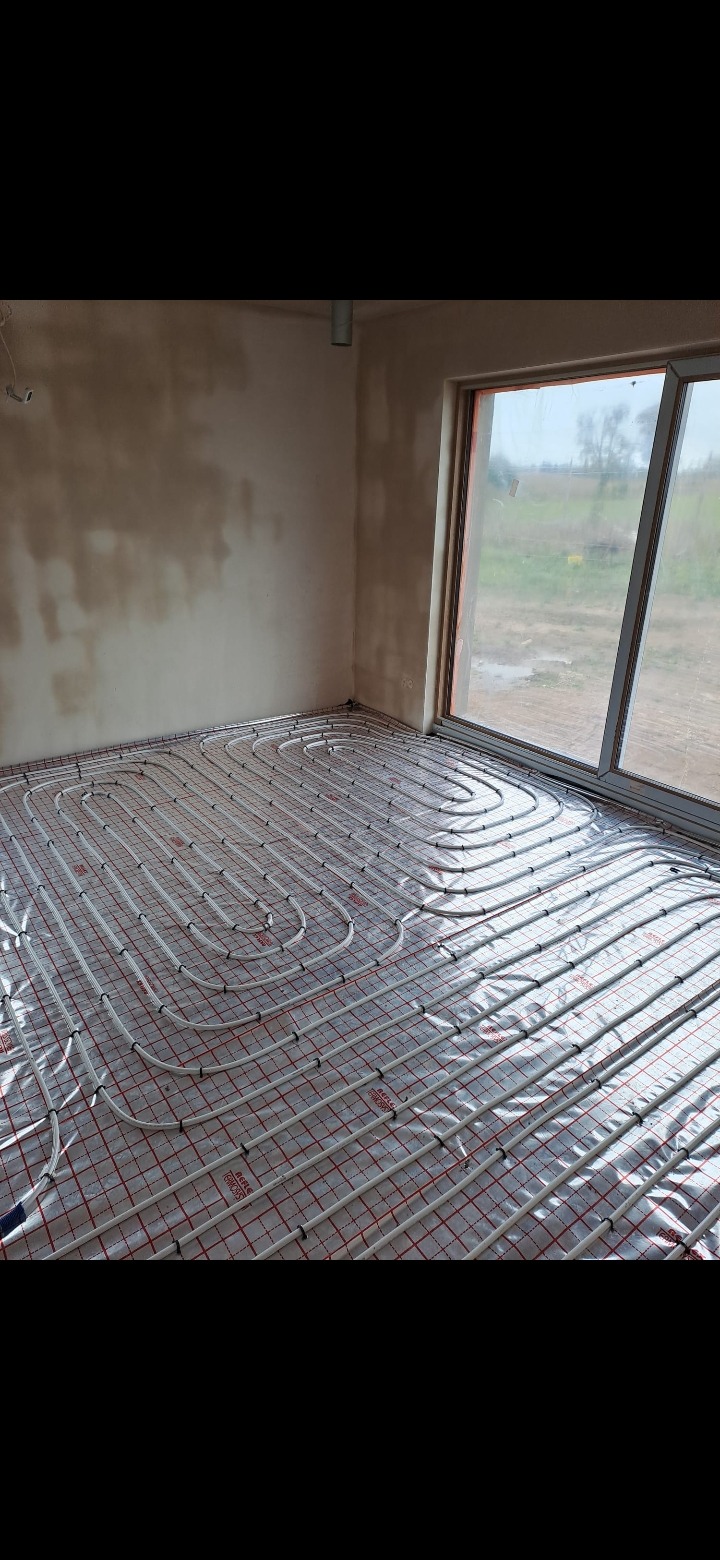 Białe rury ogrzewania podłogowego ułożone na folii z czerwoną siatką w surowym wnętrzu, widok na zewnątrz przez okno.