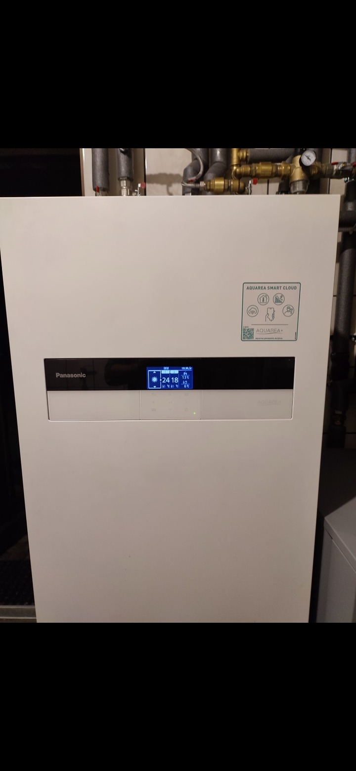 Biała jednostka wewnętrzna pompy ciepła Panasonic Aquarea Smart Cloud z widocznym wyświetlaczem LCD i naklejką informacyjną, rury instalacji grzewczej w tle.