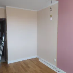 Pomalowane na beżowo ściany w pokoju z drewnianą podłogą, fragment ściany pomalowany na różowo, widoczna listwa przypodłogowa w trakcie montażu i drabina w tle.