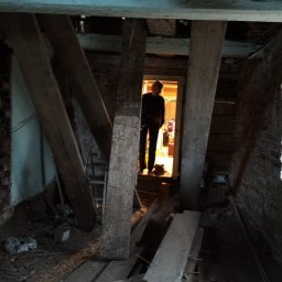 Padre złota rączka - Wnętrze poddasza w trakcie remontu, widoczne stare drewniane belki konstrukcyjne, fragment ściany z cegły i postać w przejściu do oświetlonego pomieszczenia.