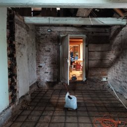 Padre złota rączka - Przygotowanie podłoża pod posadzkę w remontowanym pokoju ze starą cegłą na ścianach, widoczne zbrojenie, worek z materiałem i zwinięty przedłużacz.