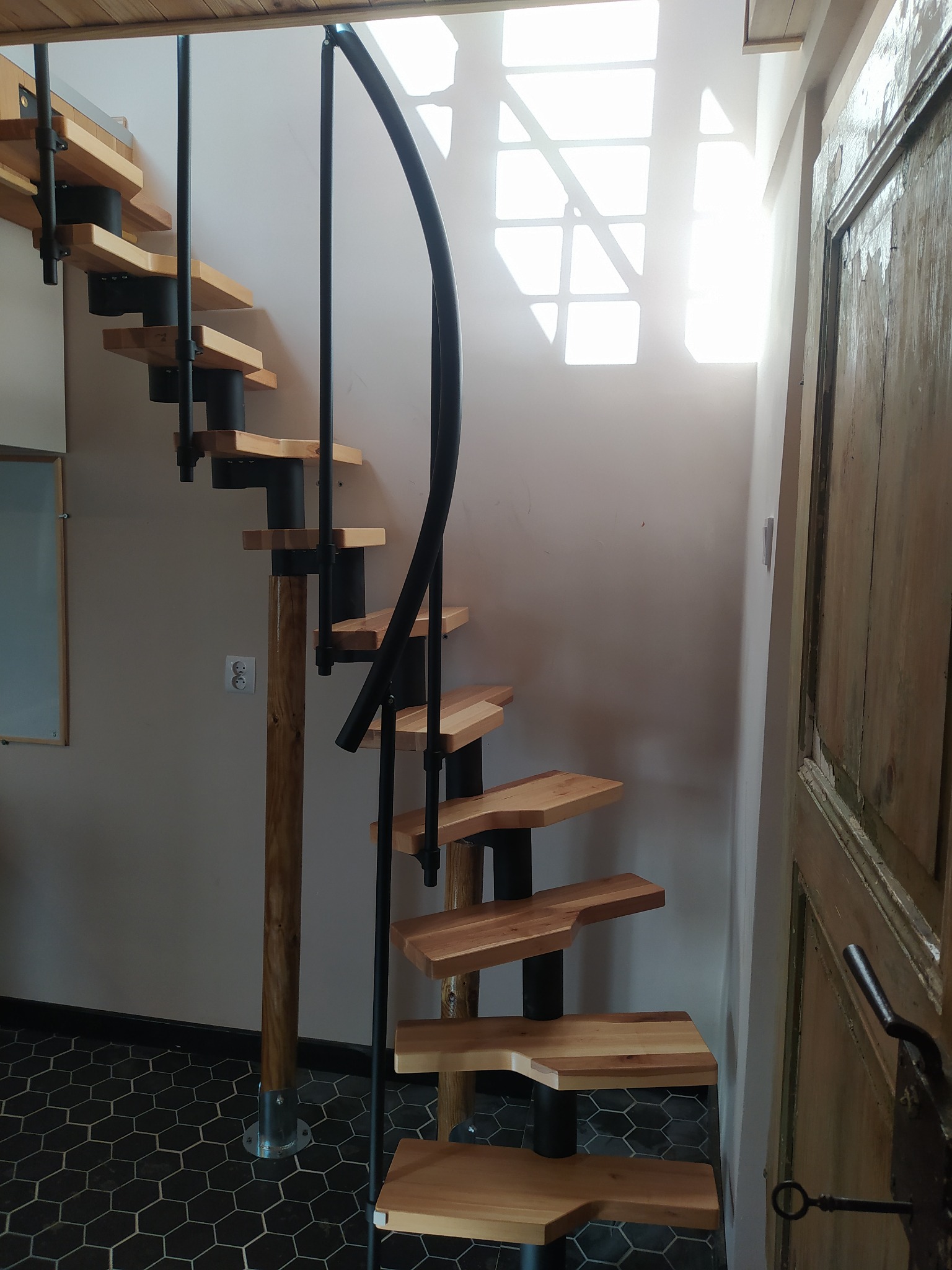 Spiralne schody z drewnianymi stopniami i czarną metalową konstrukcją, umieszczone w rogu pomieszczenia z heksagonalną podłogą i starymi, drewnianymi drzwiami po prawej stronie.