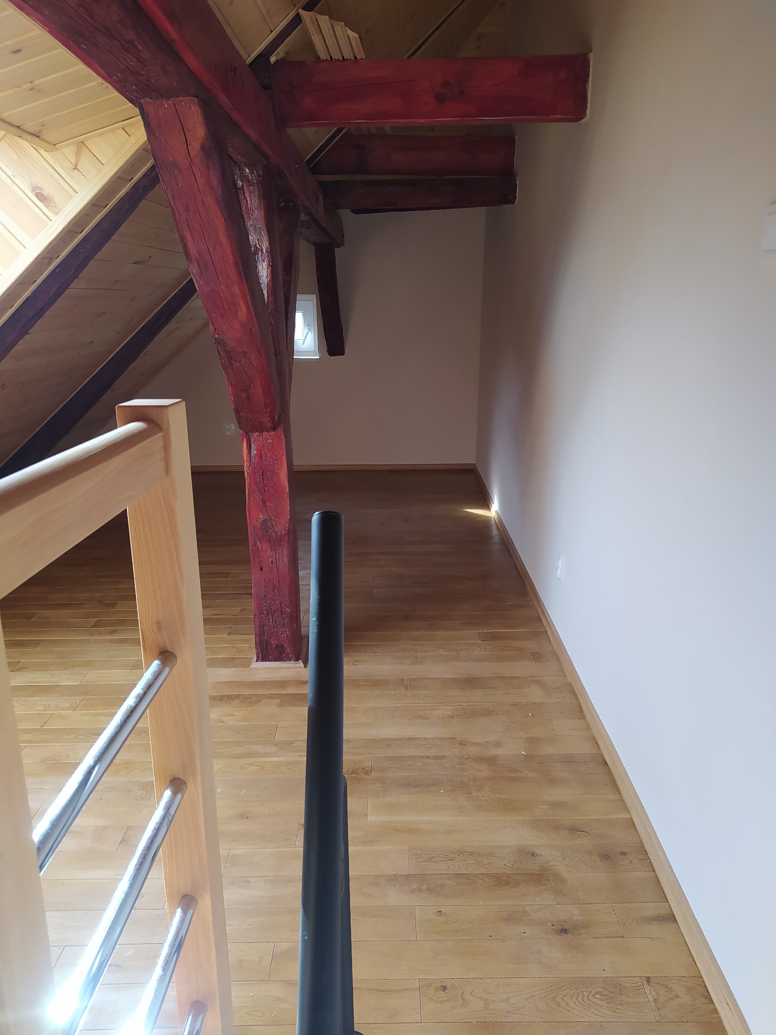 Poddasze z drewnianą podłogą, widoczne fragmenty schodów z jasnego drewna i metalowymi poręczami oraz pomalowane na czerwono drewniane elementy konstrukcyjne dachu.