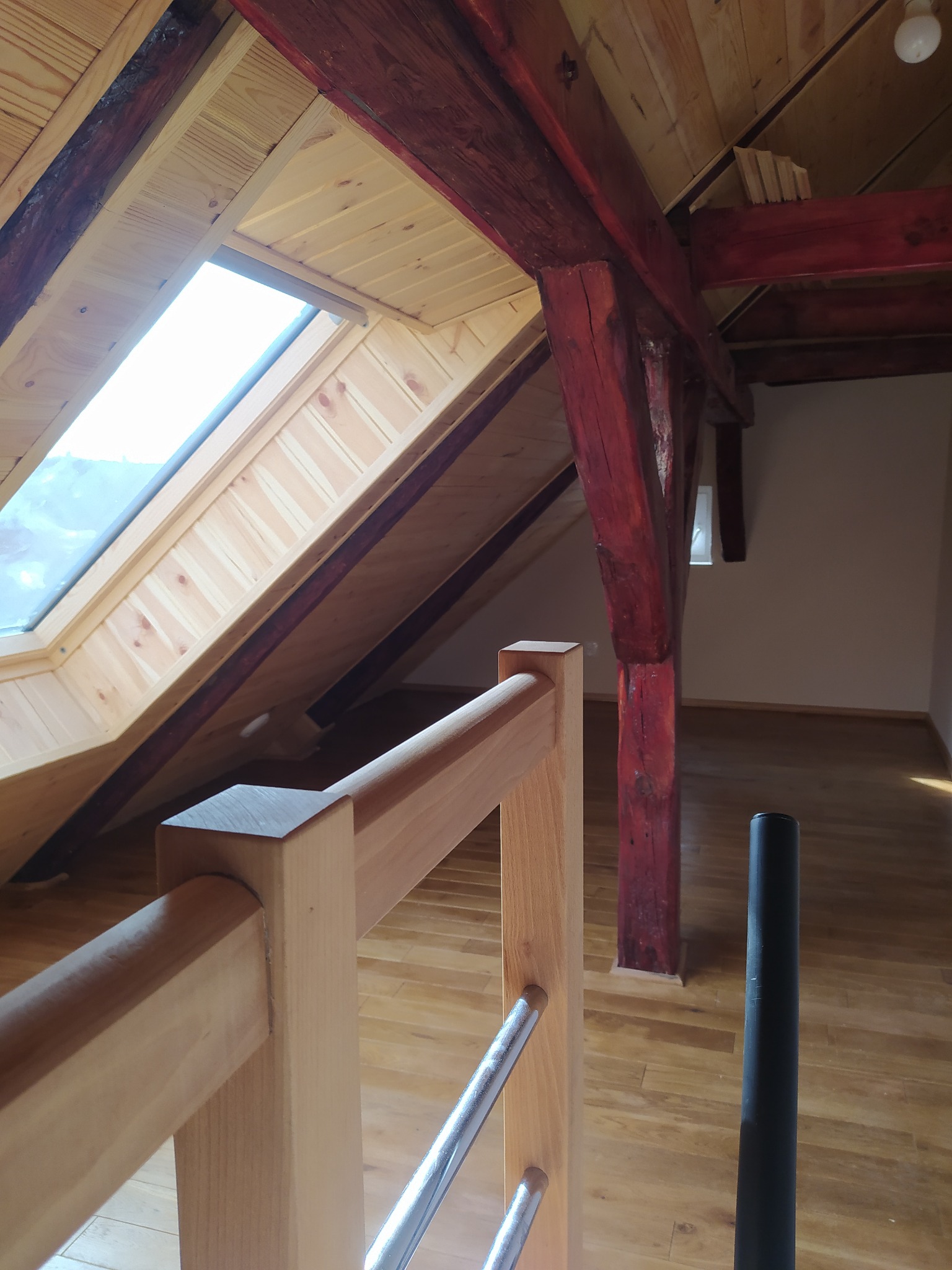 Poddasze z drewnianymi belkami pomalowanymi na czerwono, jasną drewnianą boazerią wokół okna dachowego, drewnianą podłogą i fragmentem jasnej drewnianej balustrady ze srebrnymi elementami.