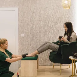 Kobieta w zielonym uniformie wykonuje zabieg na stopę klientki siedzącej w zielonym fotelu, z filiżanką w dłoni, w salonie z beżową tapetą.