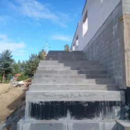 Betonowe schody prowadzące do surowego domu z bloczków, widoczne hydroizolacje fundamentów i słup energetyczny w tle.