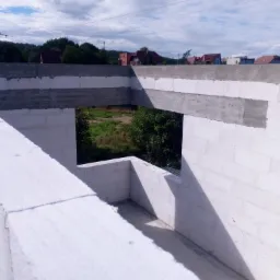 Wnętrze surowego domu w trakcie budowy z białych bloczków, widoczne betonowe nadproża i widok na zieleń za oknem.