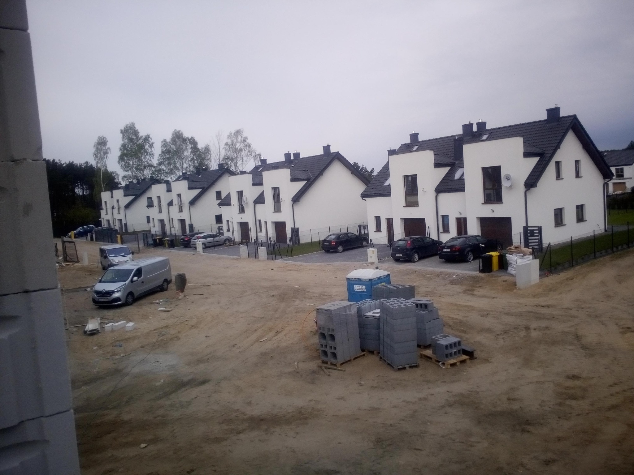 Nowo wybudowane szeregowe domy z białymi elewacjami i ciemnymi dachami, widok z placu budowy z ułożonymi bloczkami betonowymi i zaparkowanym vanem.