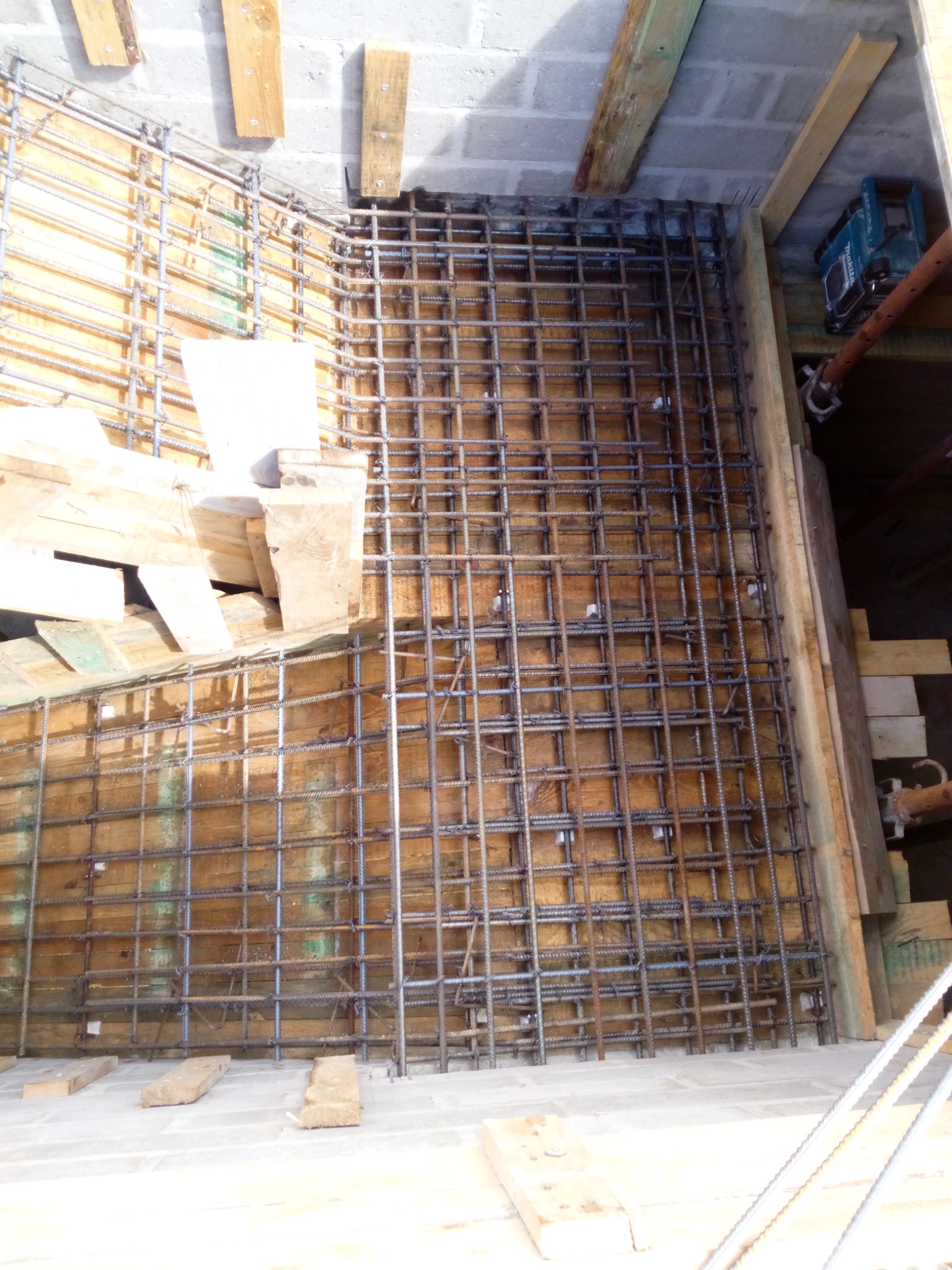 Zbrojenie fundamentu w drewnianym szalunku, widoczne pręty zbrojeniowe ułożone w kratę, drewniane deski szalunkowe i betonowe bloczki w tle.