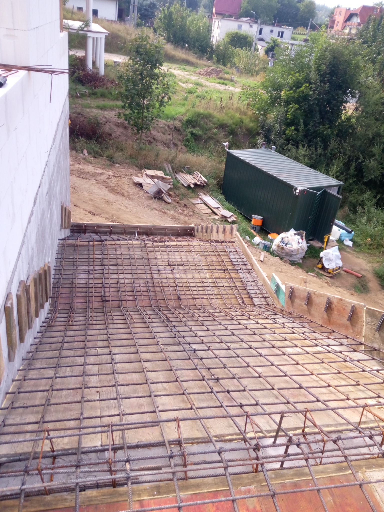 Zbrojenie fundamentu domu jednorodzinnego, widoczne szalunki i siatka z prętów stalowych, w tle kontener budowlany i częściowo wzniesiona ściana z bloczków.