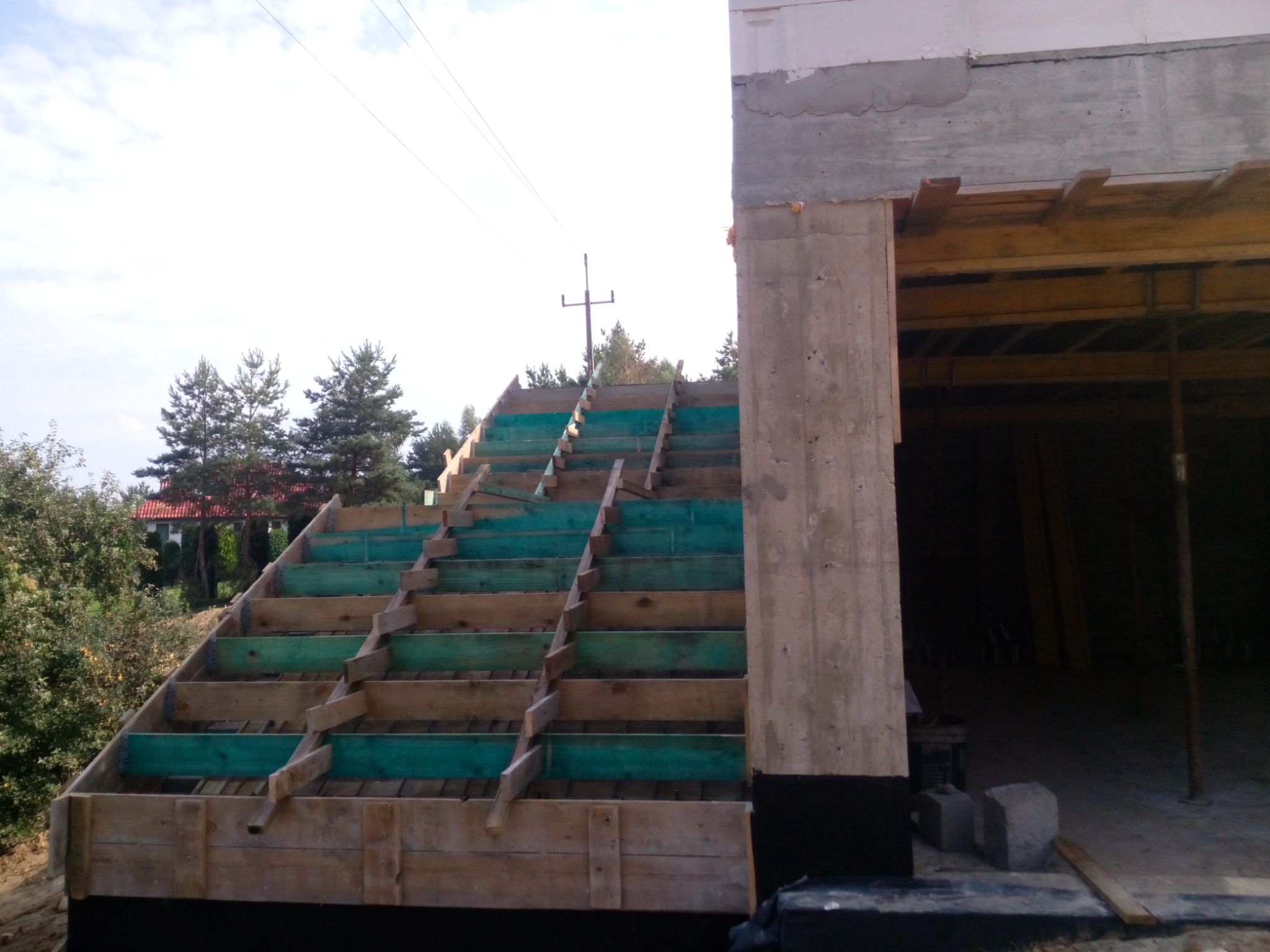 Szczegółowe ujęcie szalunku schodów zewnętrznych z zielonymi elementami, widoczne z boku budynku w trakcie budowy, na tle drzew i nieba.