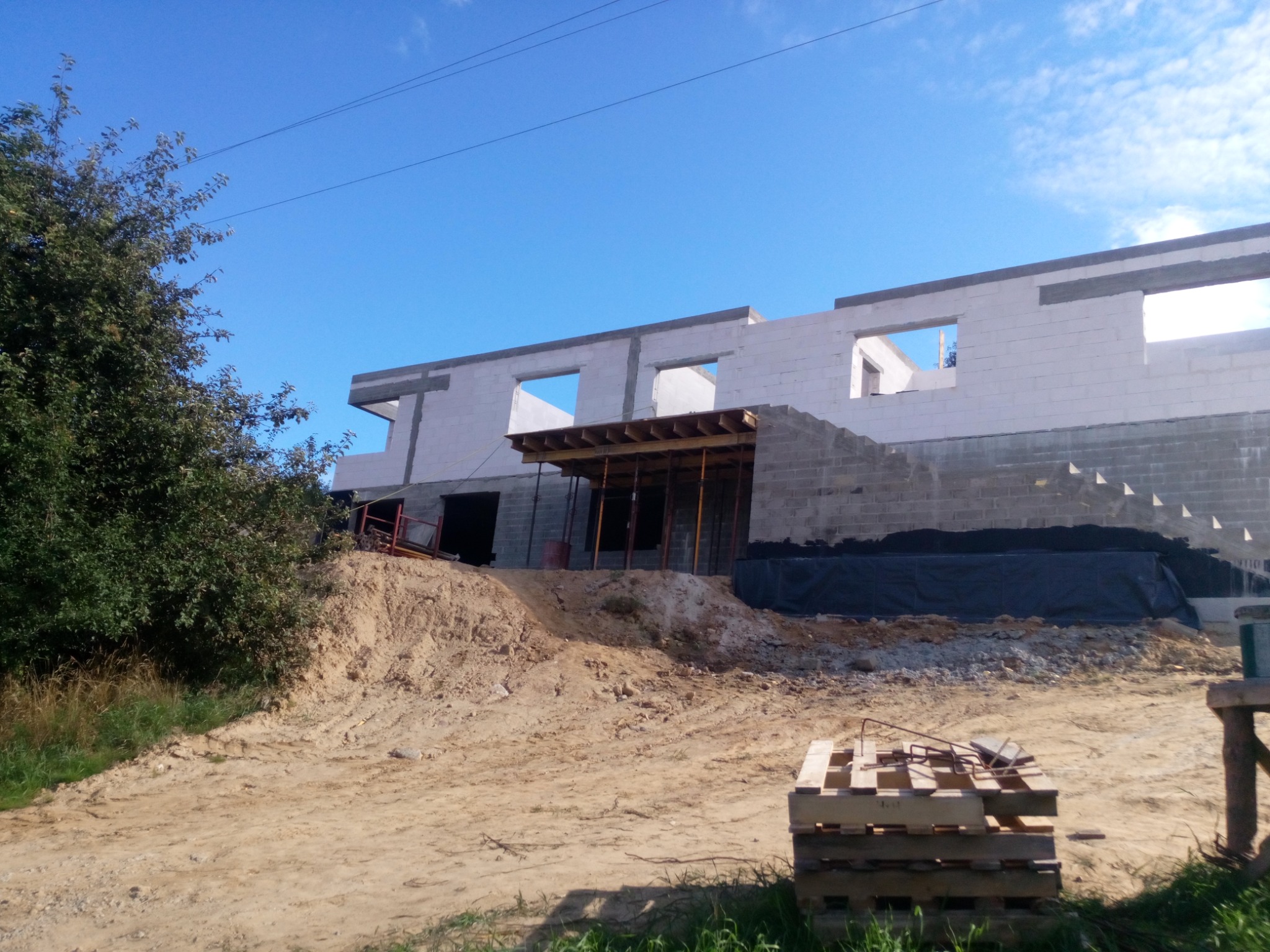 Wznoszony budynek z białych bloczków, widoczne otwory okienne i zadaszenie nad wejściem, teren budowy z ziemią i paletą.