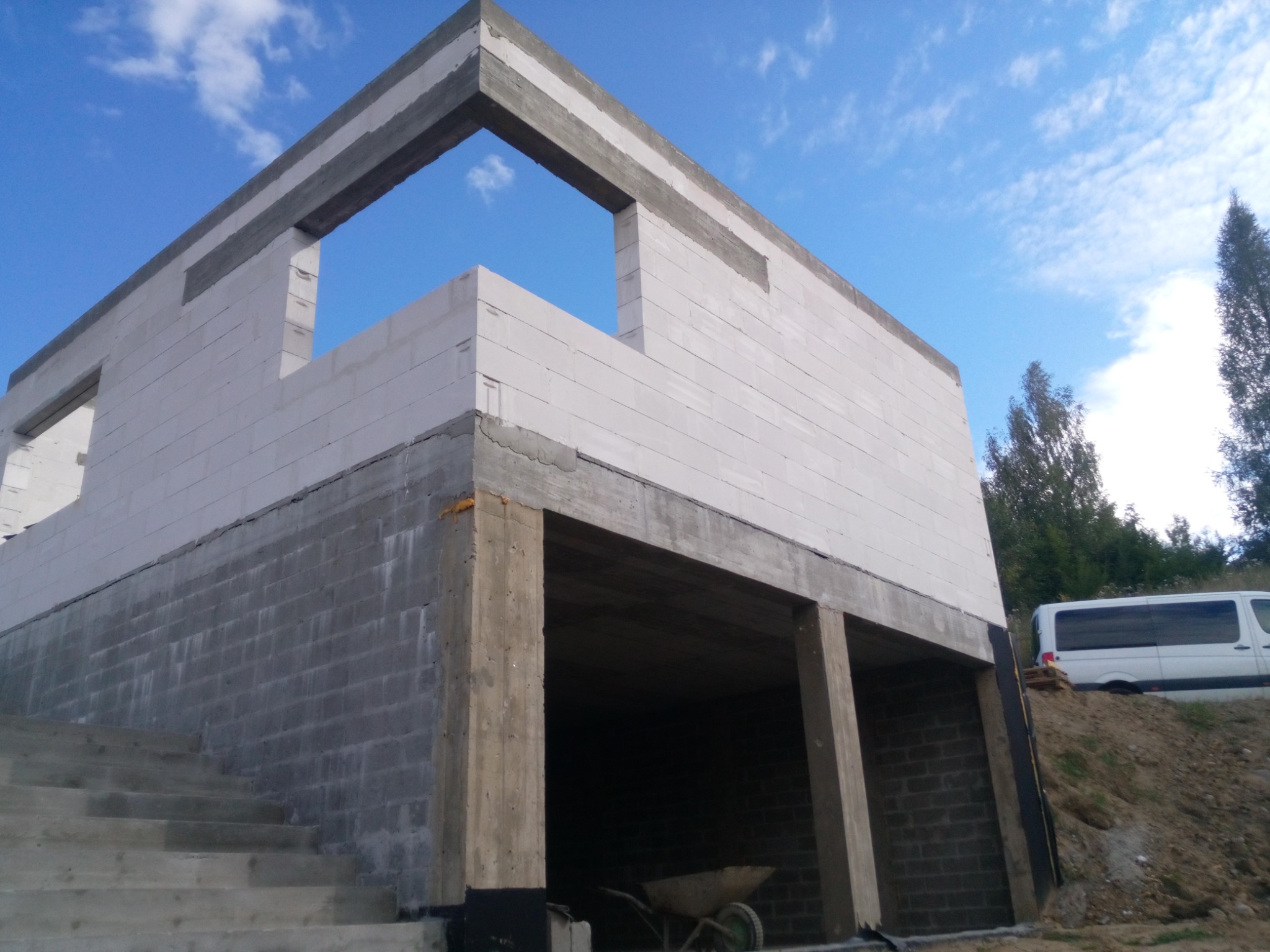 Budowa domu: ściany z białych bloczków na fundamencie z szarej cegły, widoczne betonowe filary i fragment nieba z chmurami