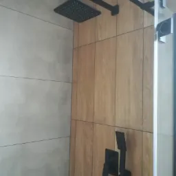 Nowoczesna kabina prysznicowa z kwadratową deszczownicą i baterią w kolorze czarnym, wykończona płytkami imitującymi drewno i szarymi płytkami.