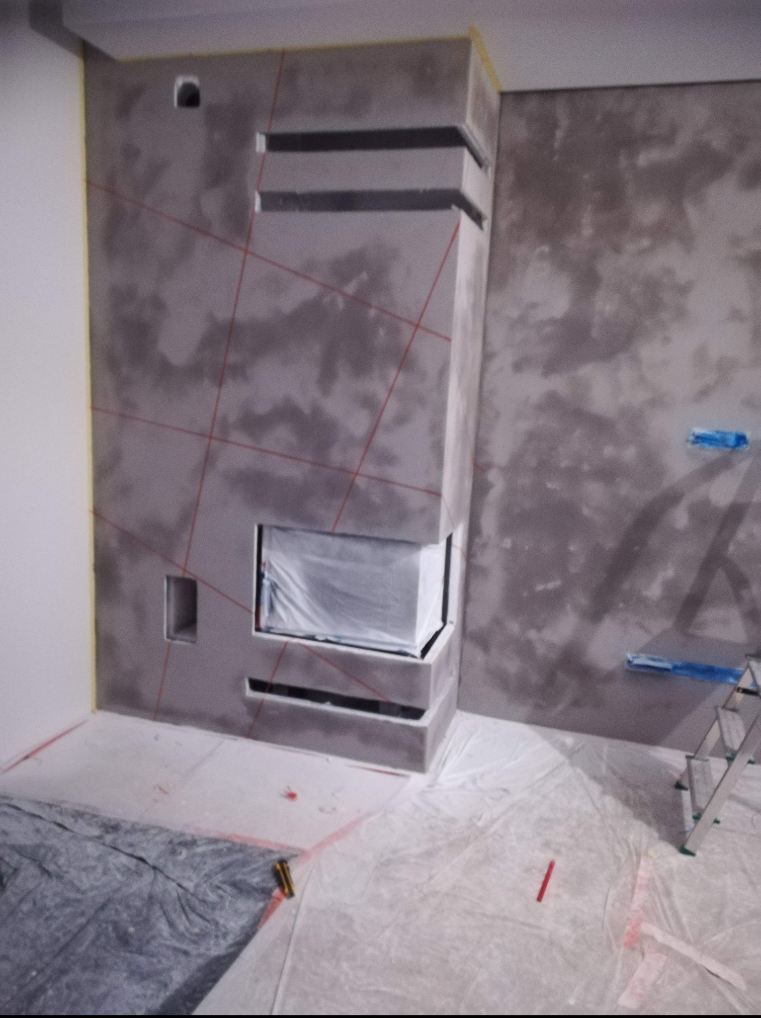 Ściana z dekoracyjnym tynkiem w odcieniach szarości, z wnęką na kominek zabezpieczoną folią, widoczne ślady po wyznaczaniu linii laserem, podłoga zabezpieczona folią malarską, drabina w rogu...