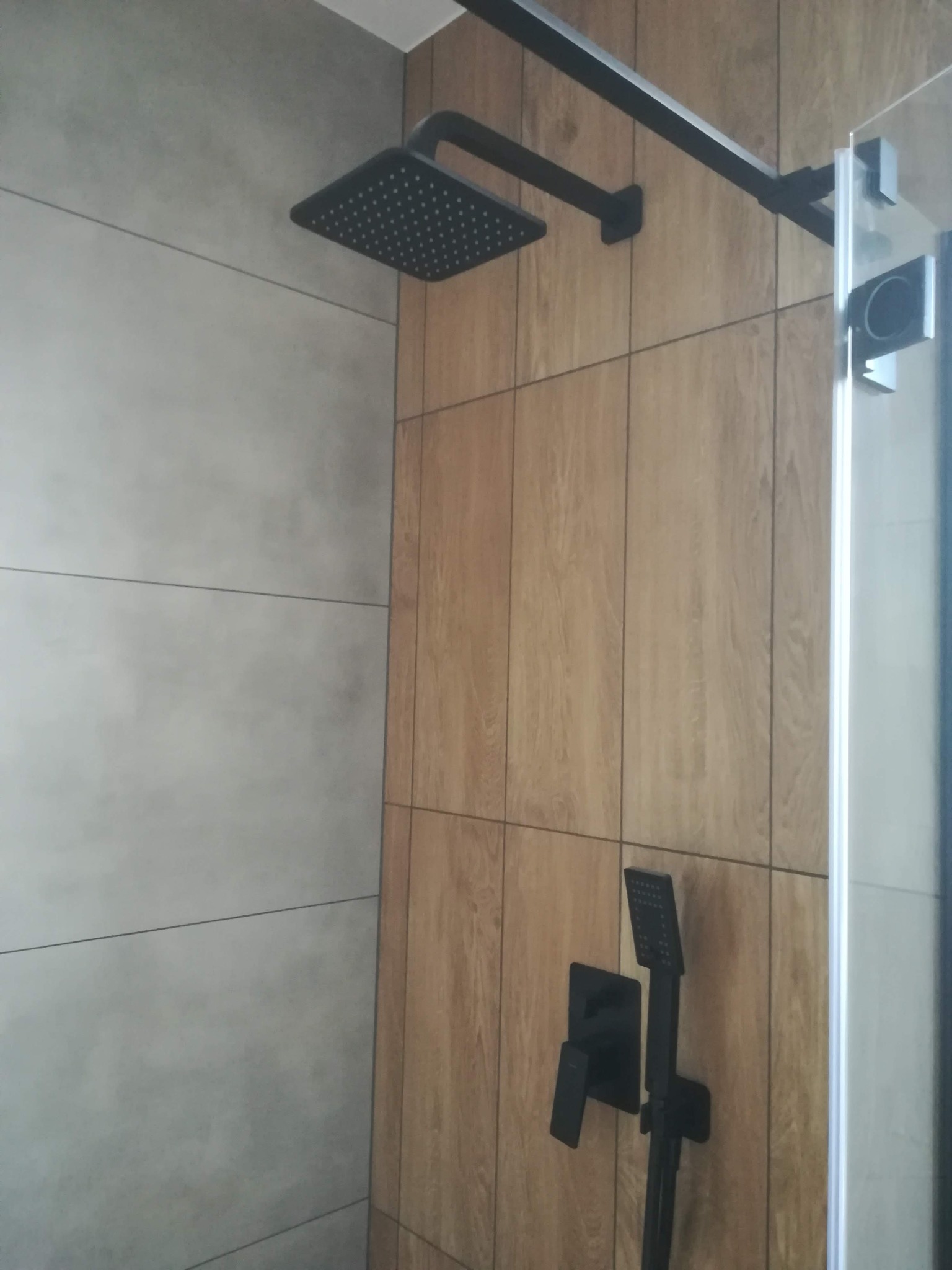Nowoczesna kabina prysznicowa z kwadratową deszczownicą i baterią w kolorze czarnym, wykończona płytkami imitującymi drewno i szarymi płytkami.