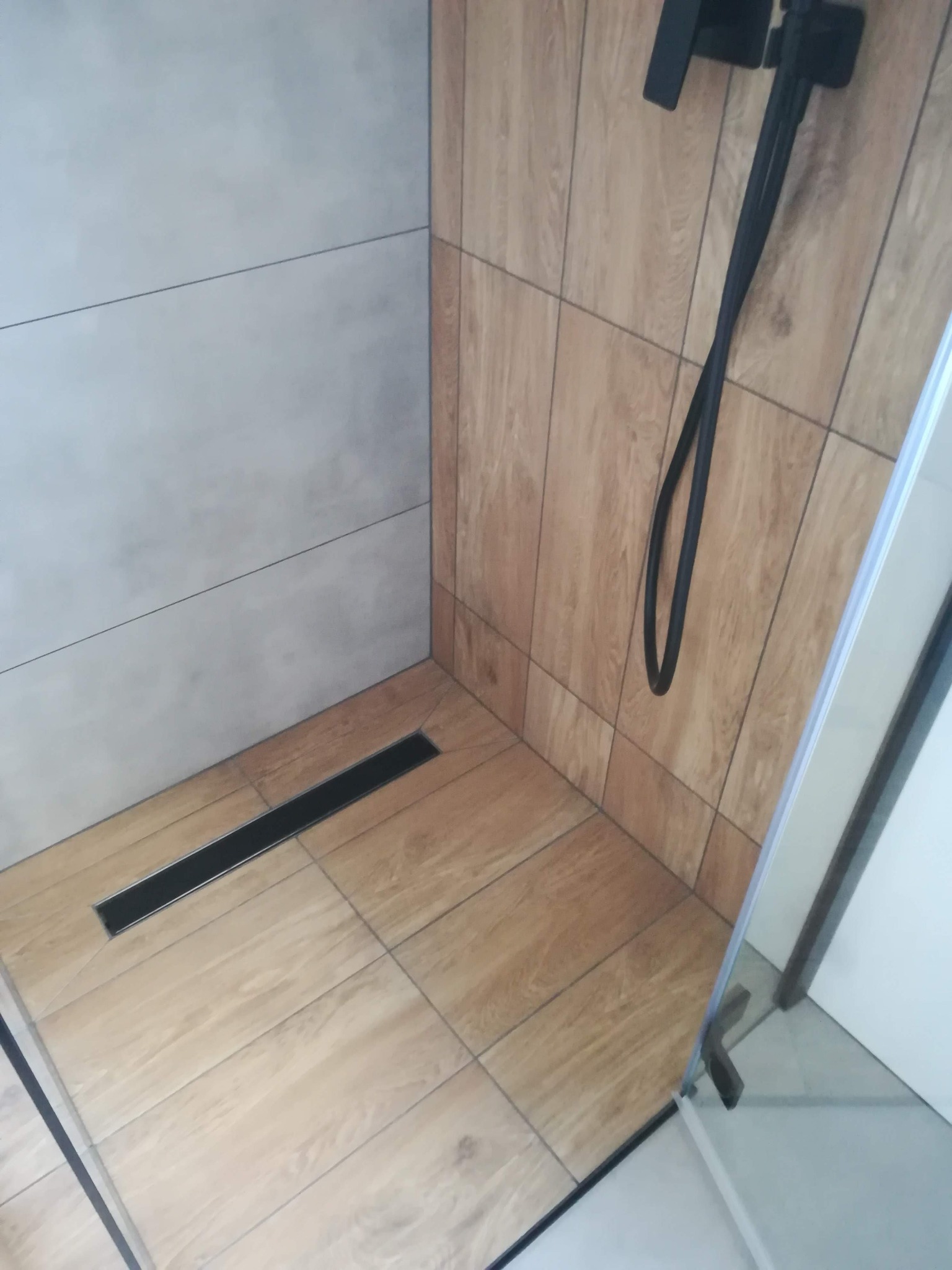 Kabina prysznicowa z odpływem liniowym, wykończona płytkami imitującymi drewno na ścianie i podłodze, oraz szarymi płytkami na przeciwległej ścianie, widoczny czarny zestaw prysznicowy.